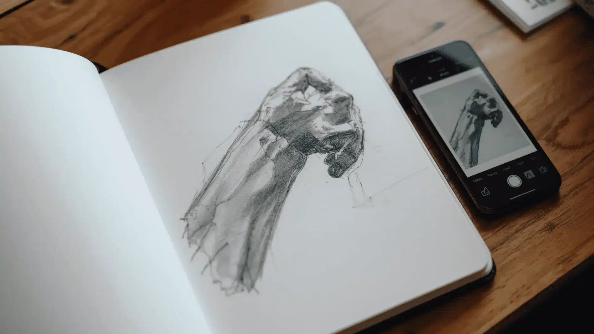 Foreshortened hand study in sketchbook with closer parts larger, reference image visible on a phone