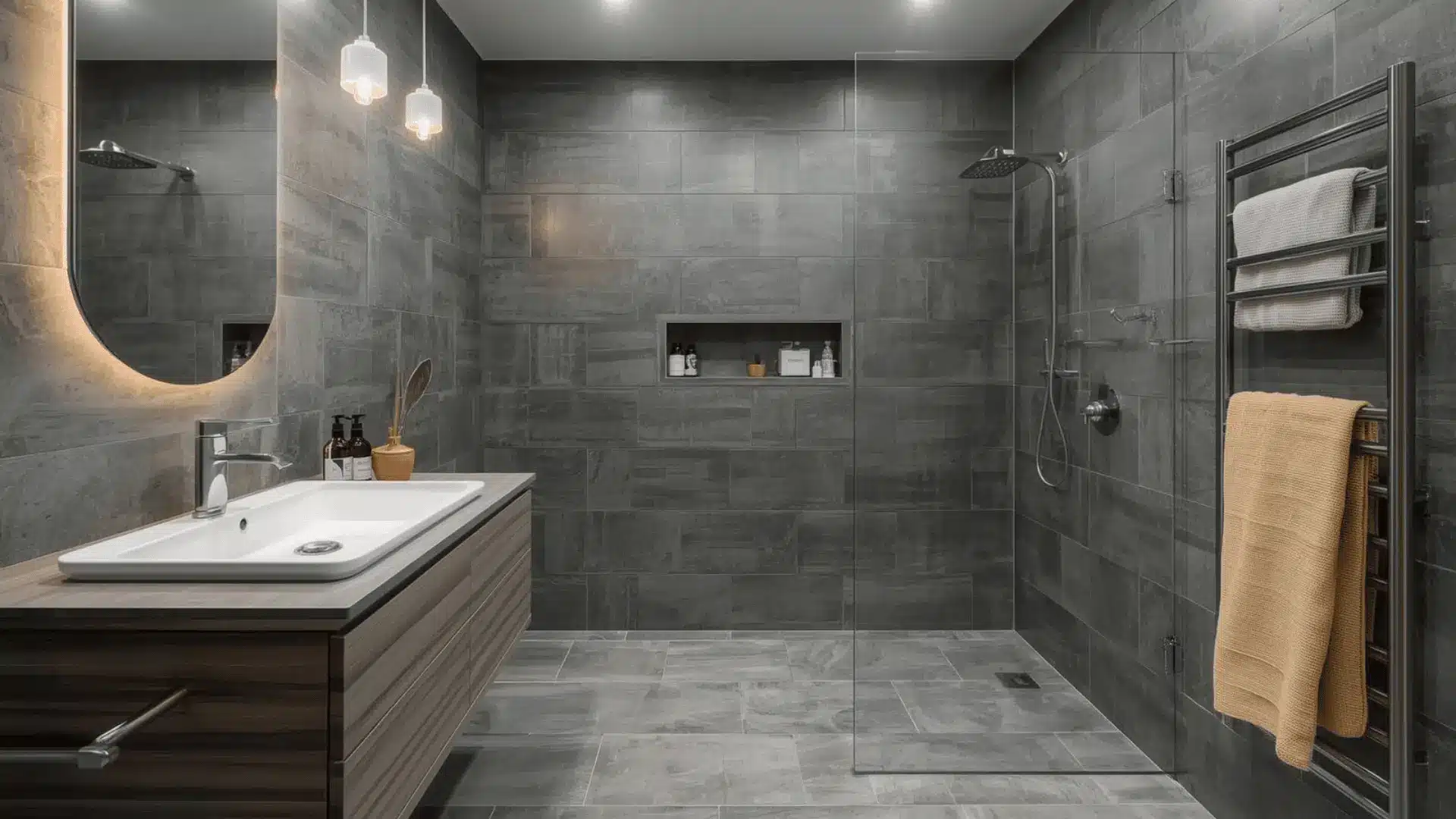 Modern wet room with dark grey tiles, open shower zone, frameless glass panel, niche shelf, and backlit mirror.