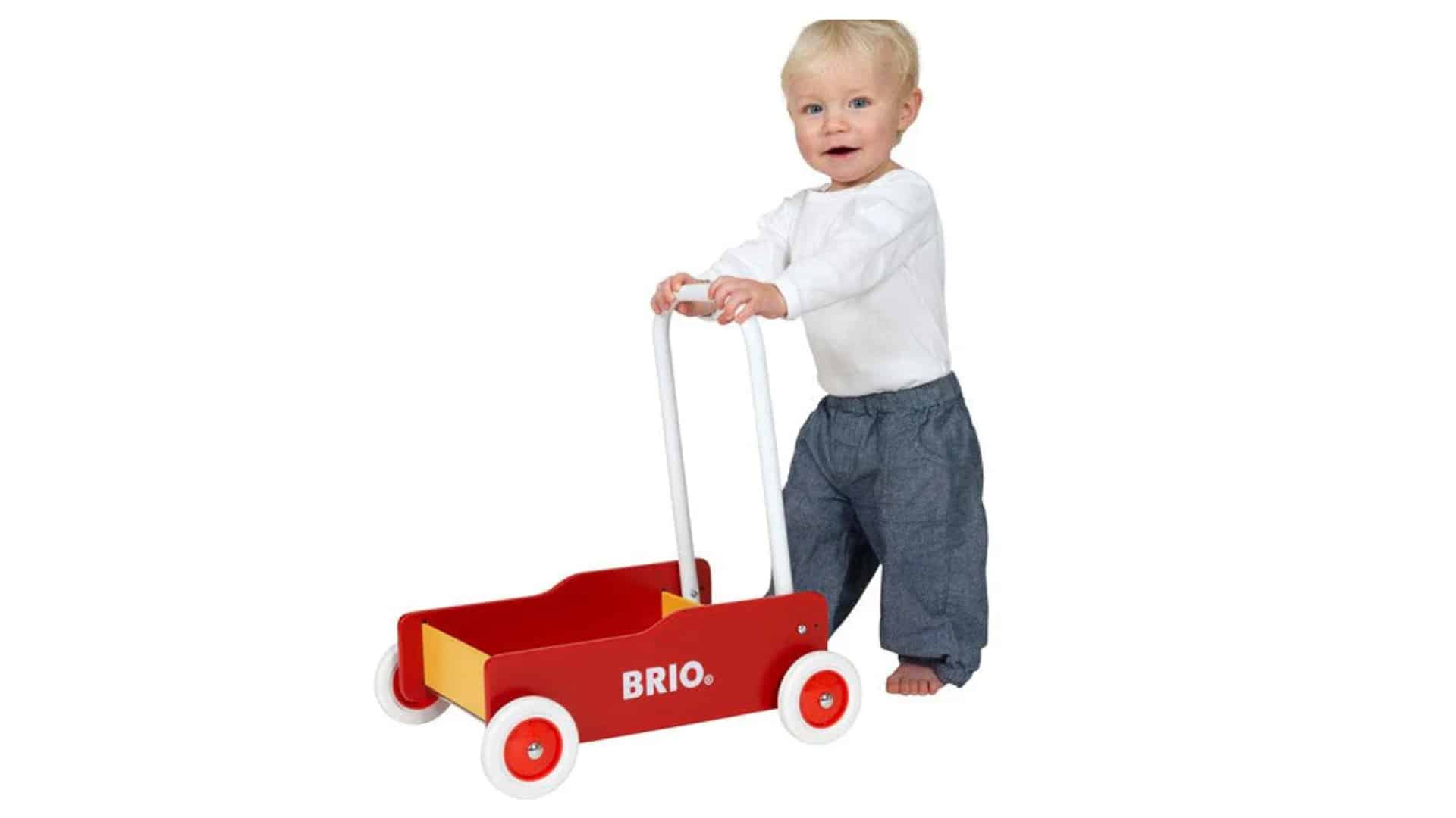 Red Brio toddler walker cart with white handle and wheels, shown beside a standing baby in a clean white studio photo.
