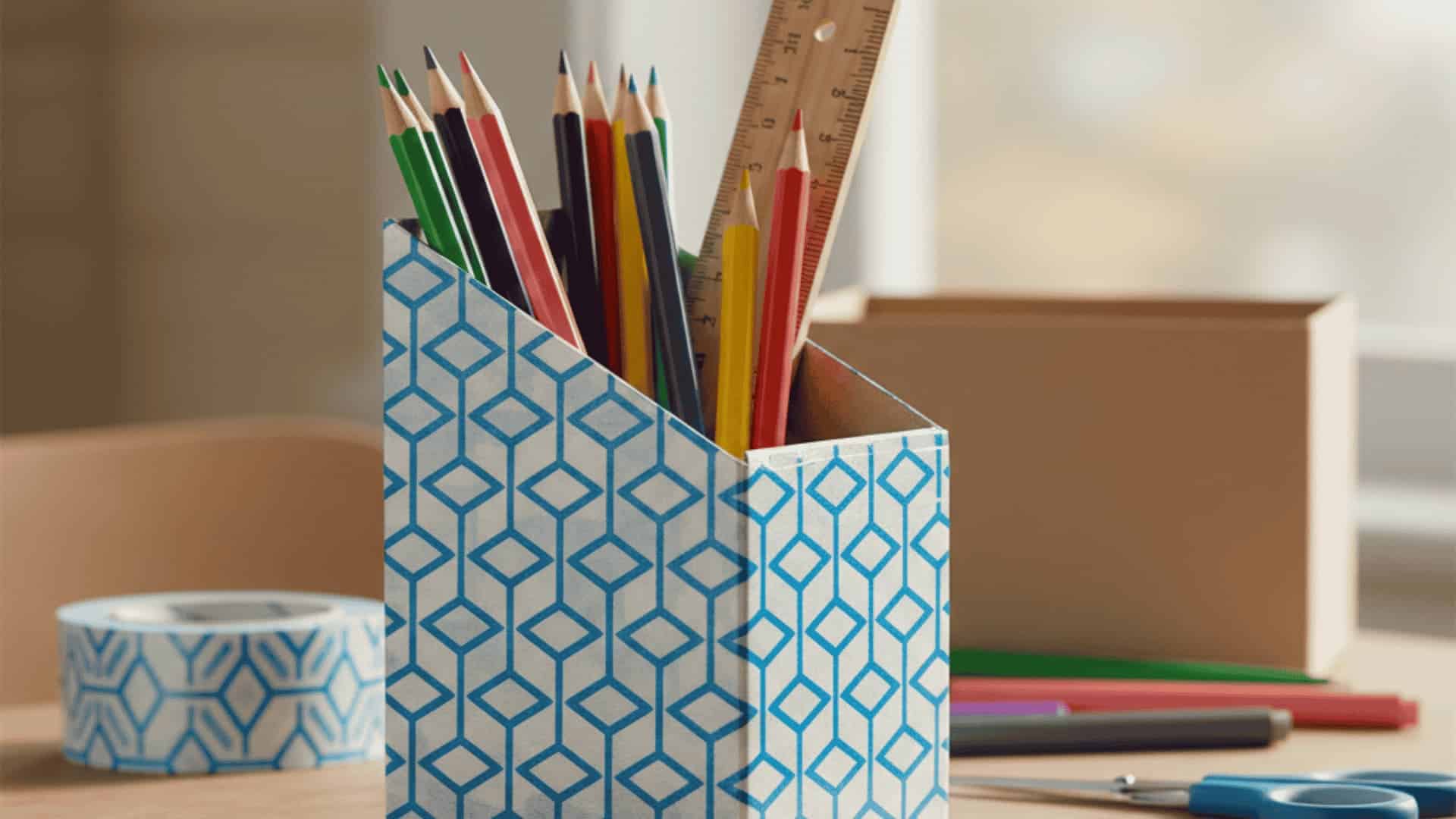 Duct tape decorated cereal box organizer holding colored pencils on wooden desk with tape roll and markers nearby
