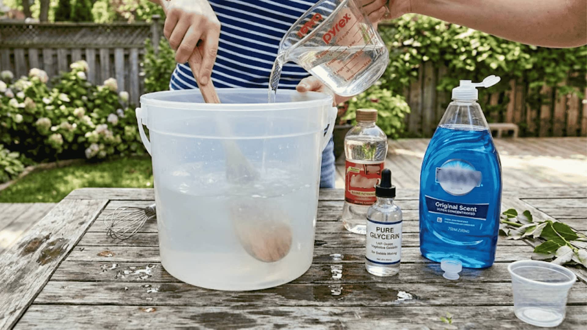 Person pouring water into a bucket while mixing homemade giant bubble solution with glycerin and dish soap on an outdoor table