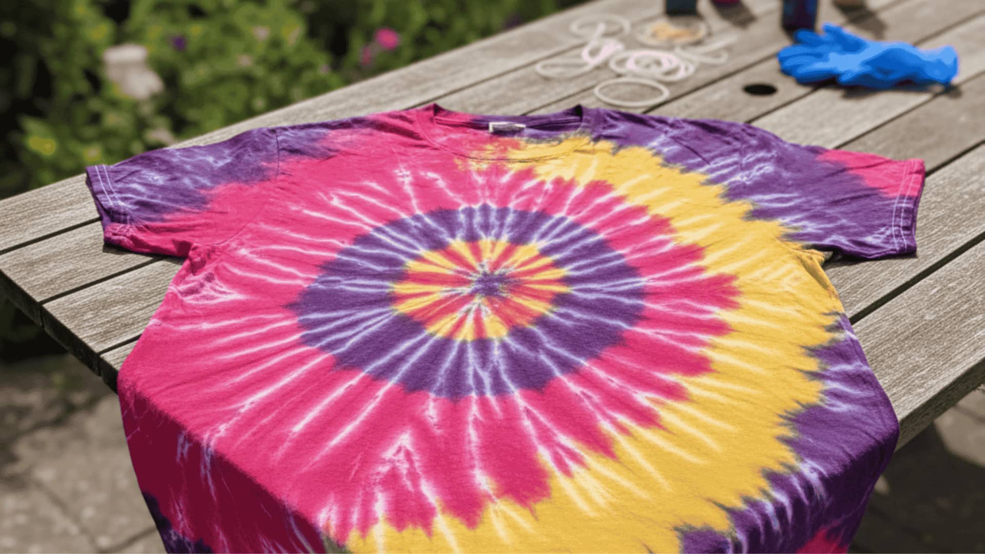 Pink, purple, and yellow spiral tie-dye t-shirt laid flat on a garden table with dye bottles and rubber bands nearby
