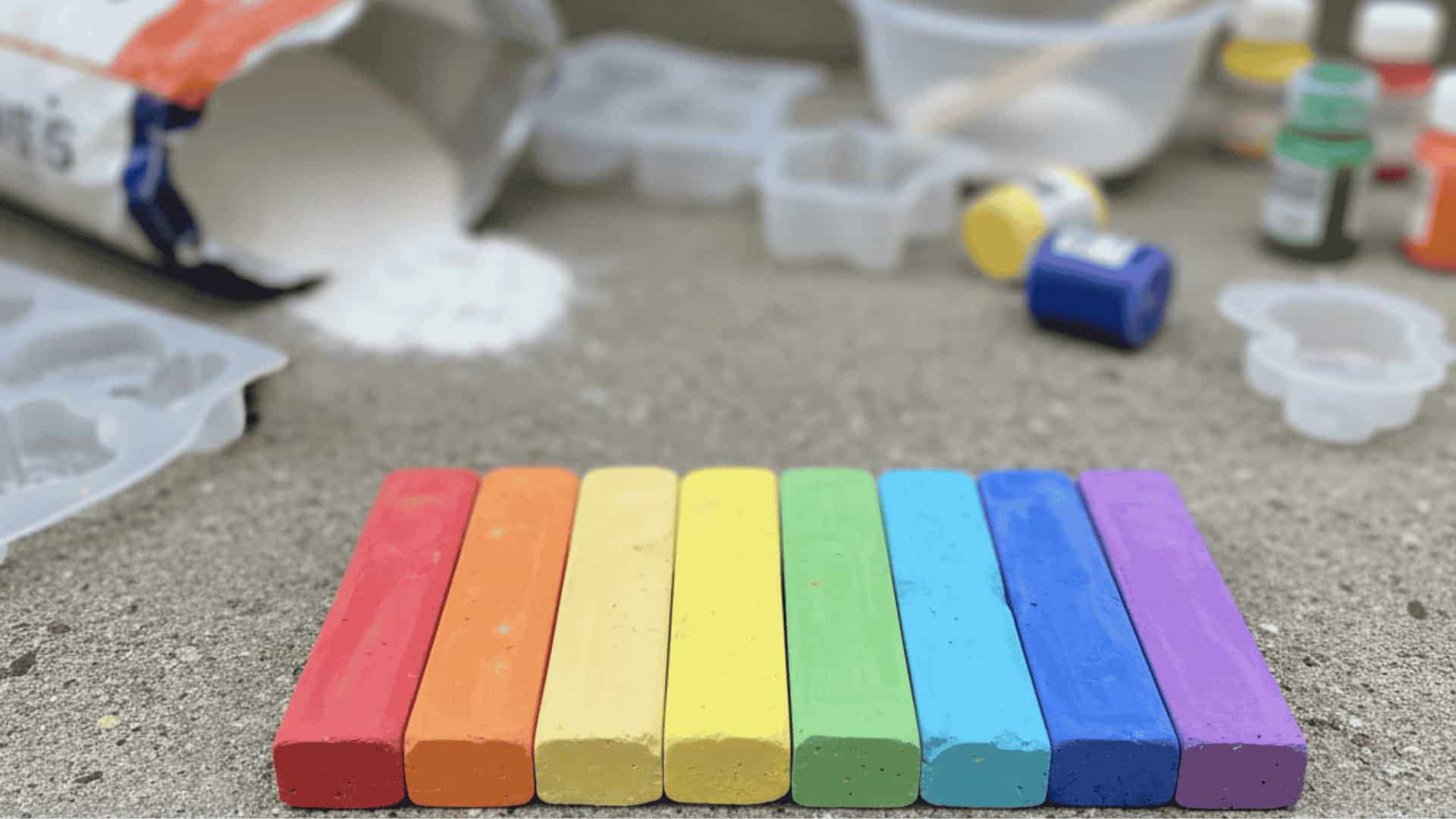 Six rainbow-colored homemade chalk sticks on a concrete step with plaster bag silicone molds and tempera paint bottles