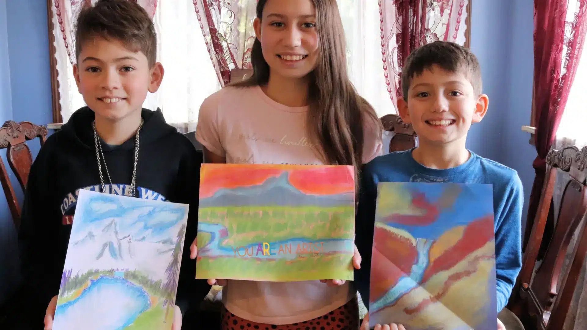 Three smiling kids hold colorful chalk pastel landscape artworks indoors, showcasing the “You Are An Artist” fine arts program.
