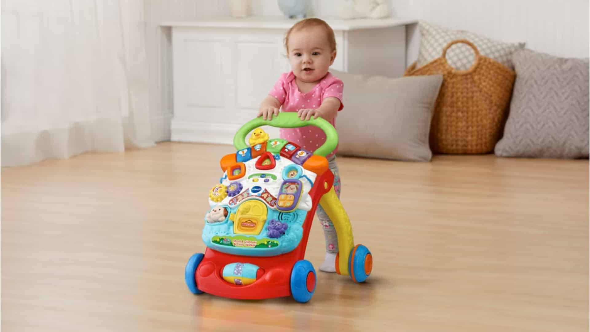 Baby standing and pushing a VTech sit-to-stand walker with a colorful activity panel in a bright living room setting. For play.